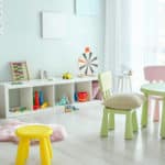 stanzetta di asilo nido con tavolini e sgabelli da bimbi colorati sulla parete in fondo degli scaffali con giochi e un pouff sullo sfondo una porta finestra con tende bianche