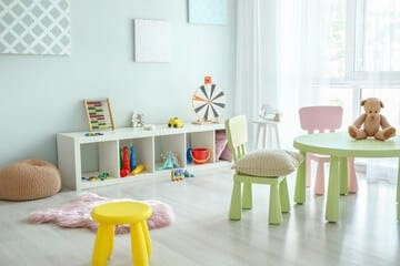 stanzetta di asilo nido con tavolini e sgabelli da bimbi colorati sulla parete in fondo degli scaffali con giochi e un pouff sullo sfondo una porta finestra con tende bianche