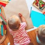 inquadratura dall'alto di 2 bimbi piccolini che giocano sul parquet con dei colori e fogli