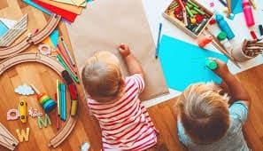 inquadratura dall'alto di 2 bimbi piccolini che giocano sul parquet con dei colori e fogli